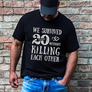 K&ouml;nnte beinhalten: Schwarzes T-Shirt mit wei&szlig;em Text, der "We survived 20 years without killing each other" lautet. Der Text ist in einer stilisierten Schriftart mit einem Herzsymbol.