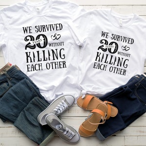 K&ouml;nnte beinhalten: Zwei wei&szlig;e T-Shirts mit schwarzem Text, der "Wir haben 20 Jahre &uuml;berlebt, ohne uns zu t&ouml;ten" lautet. Die Shirts liegen auf einer Holzoberfl&auml;che mit einer blauen Jeans und einem Paar Sandalen.