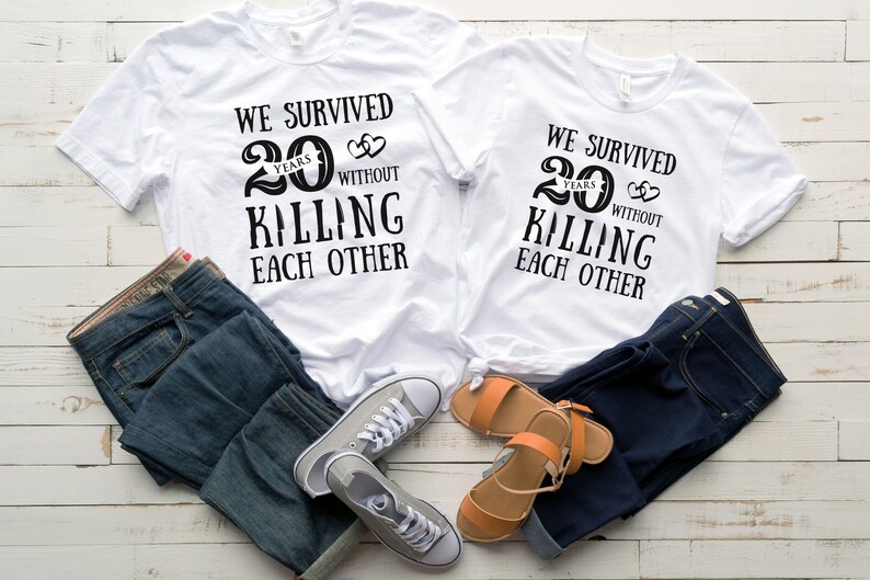 K&ouml;nnte beinhalten: Zwei wei&szlig;e T-Shirts mit schwarzem Text, der "Wir haben 20 Jahre &uuml;berlebt, ohne uns zu t&ouml;ten" lautet. Die Shirts liegen auf einer Holzoberfl&auml;che mit einer blauen Jeans und einem Paar Sandalen.