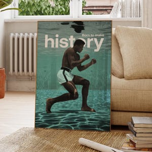 Può includere: Una stampa artistica incorniciata con un pugile sott'acqua. L'immagine ha uno sfondo verde acqua con la parola "history" e la frase "Born to make" stampate in bianco. Il pugile indossa pantaloncini bianchi con una striscia nera.