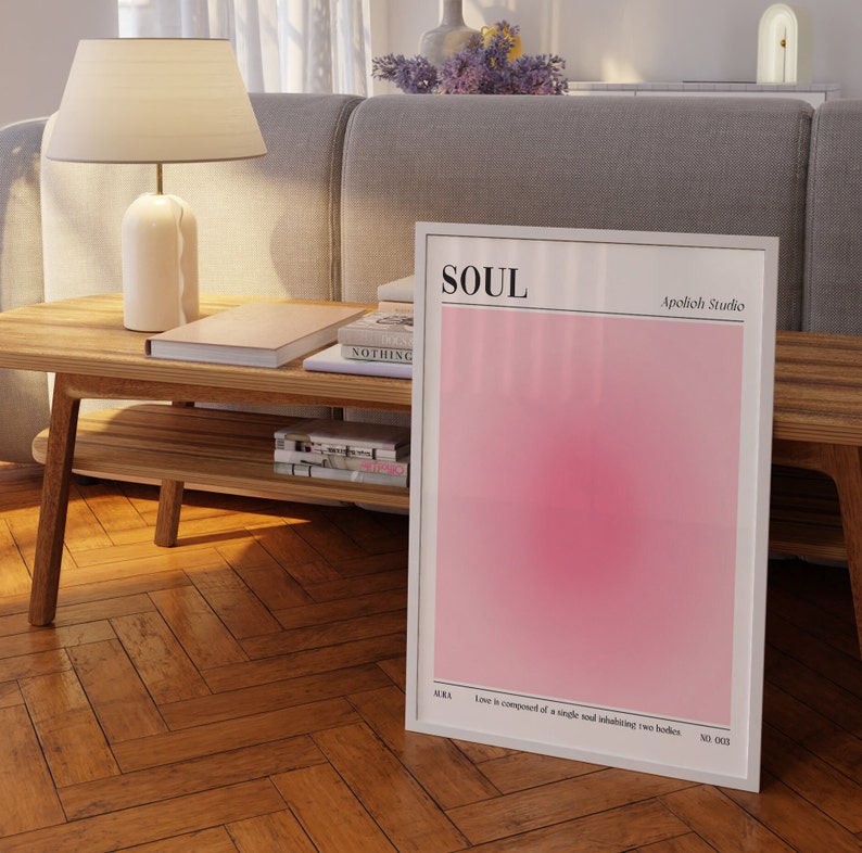 May include: A framed art print with the word "SOUL" at the top and "Apolioh Studio" below it. The print features a gradient of pink hues. The text "AURA" and a quote are at the bottom. The print is displayed on a wooden table.