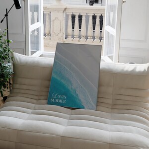 K&ouml;nnte beinhalten: Ein wei&szlig;es Sofa mit einem gerahmten Druck einer Luftaufnahme von t&uuml;rkisfarbenen Ozeanwellen, die an einem wei&szlig;en Sandstrand brechen. Der Druck tr&auml;gt den Text "Lovin Summer".