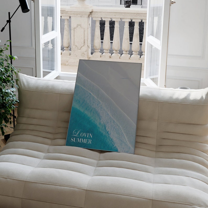 K&ouml;nnte beinhalten: Ein wei&szlig;es Sofa mit einem gerahmten Druck einer Luftaufnahme von t&uuml;rkisfarbenen Ozeanwellen, die an einem wei&szlig;en Sandstrand brechen. Der Druck tr&auml;gt den Text "Lovin Summer".