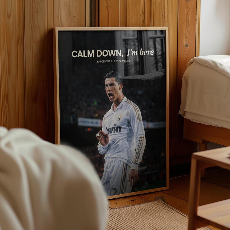 May include: A framed poster with a black and white image of a footballer in a white kit. The poster has the text "CALM DOWN, I'm here" and "BARCELONA 1-2 REAL MADRID". The frame is made of wood.