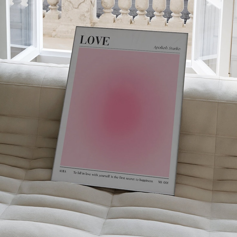 May include: A framed poster with the word "LOVE" at the top. The poster has a pink gradient background with the text "Apolioh Studio" and "AURA To fall in love with yourself is the first secret to happiness NO.001". The poster is leaning against a beige sofa.