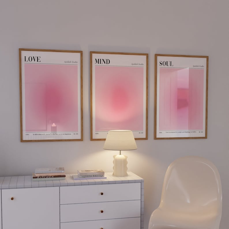 May include: Three framed art prints with the words "LOVE", "MIND", and "SOUL" in a minimalist style. Each print features a gradient of pink hues. A white lamp and a white dresser with gold knobs are in the foreground.