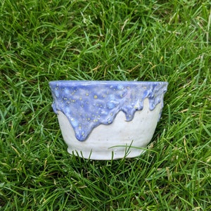 May include: A handmade ceramic bowl with a white base and a blue, drippy glaze. The blue glaze has yellow speckles and drips down the sides of the bowl. The bowl is sitting on a bed of green grass.