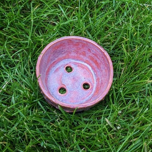 May include: A small, terracotta-colored ceramic pot with four drainage holes. The pot has a speckled, reddish-brown glaze.