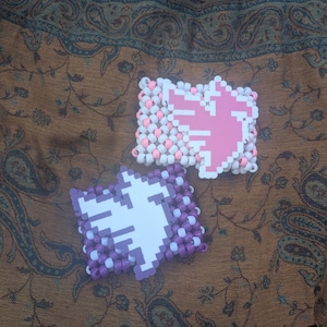 May include: Two handmade beaded bracelets. One features a pink pixelated design on a white background, surrounded by white and pink beads. The other has a white pixelated design on a purple background, surrounded by purple and white beads.