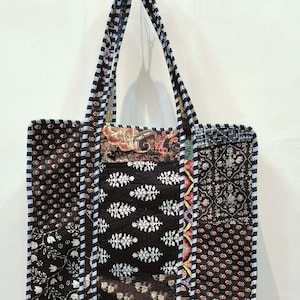 以下が含まれることがあります： 黒、白、赤、青の様々な柄の生地を使ったパッチワークのトートバッグ。バッグは長方形で、ストライプの縁とマッチしたハンドルが付いています。バッグは物を運ぶように設計されています。