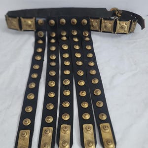 Puede incluir: Un cinturón de cuero negro con detalles en metal dorado y cinco correas colgantes con tachuelas de metal dorado. El cinturón es una réplica de un cinturón militar romano.