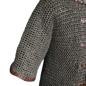 May include: A close-up of a chainmail shirt with brown leather trim. The shirt is made of interlocking metal rings and has a textured surface.