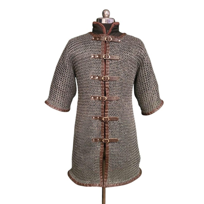 May include: A gray chainmail shirt with brown leather straps and buckles. The shirt has short sleeves and a long torso. It is displayed on a mannequin.