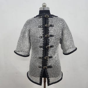 May include: A silver chainmail shirt with short sleeves, displayed on a mannequin. The shirt features black leather accents along the front and edges, secured with multiple buckles. The garment is designed for historical reenactment or costume use.