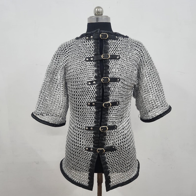 May include: A silver chainmail shirt with short sleeves, displayed on a mannequin. The shirt features black leather accents along the front and edges, secured with multiple buckles. The garment is designed for historical reenactment or costume use.