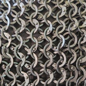 Op de afbeelding: Een close-up van een metalen mali&euml;nkolderstof met in elkaar grijpende cirkelvormige ringen in een donker gunmetal grijze afwerking. Elke individuele ring is vastgezet met een kleine klinknagel, wat een dicht, herhalend gaaspatroon op donkerbruine stof vormt.