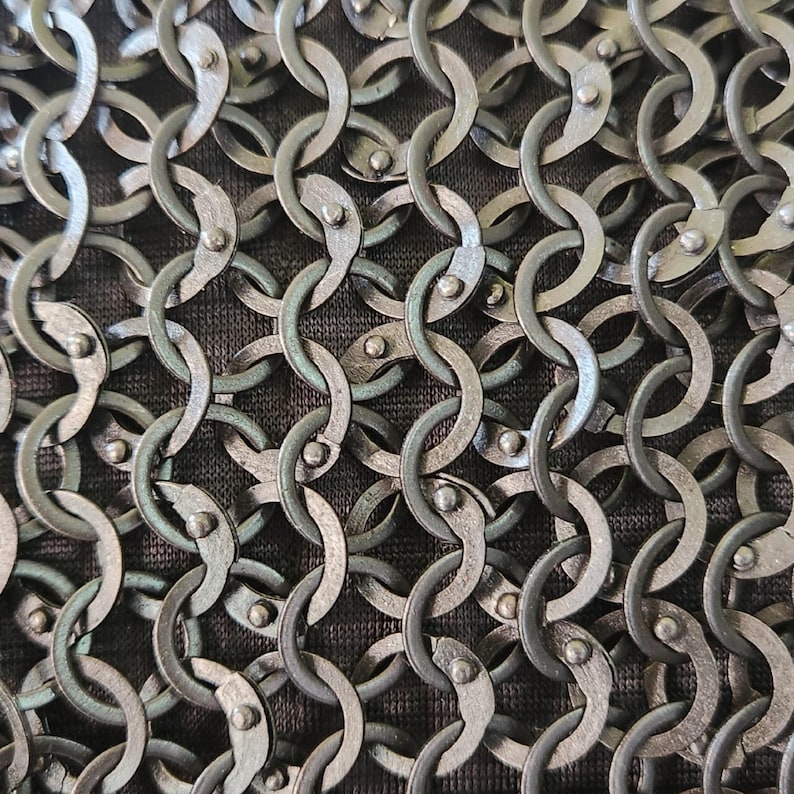 Op de afbeelding: Een close-up van een metalen mali&euml;nkolderstof met in elkaar grijpende cirkelvormige ringen in een donker gunmetal grijze afwerking. Elke individuele ring is vastgezet met een kleine klinknagel, wat een dicht, herhalend gaaspatroon op donkerbruine stof vormt.