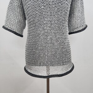 May include: A silver chainmail shirt with short sleeves and a rounded neckline. The edges of the sleeves and the bottom hem are trimmed with black material. The shirt is displayed on a stand.
