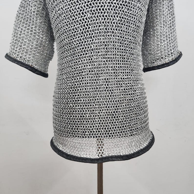 May include: A silver chainmail shirt with short sleeves and a rounded neckline. The edges of the sleeves and the bottom hem are trimmed with black material. The shirt is displayed on a stand.