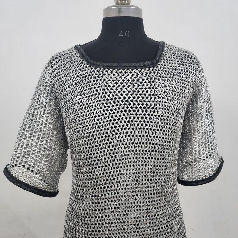 May include: A silver chainmail shirt with short sleeves and a square neckline. The edges of the sleeves and neckline are trimmed with black material. The shirt is displayed on a black mannequin.
