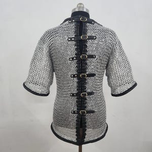 May include: A silver chainmail shirt with short sleeves and black leather accents. The back features a vertical strip of black leather with multiple buckle closures. The sleeves and neckline are trimmed with black leather.