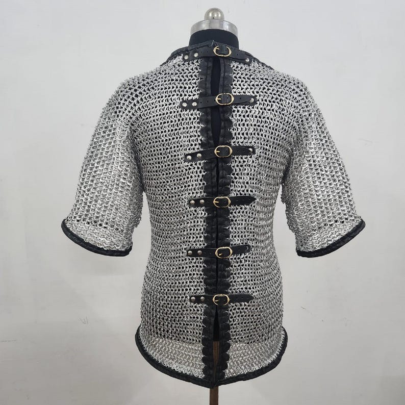 May include: A silver chainmail shirt with short sleeves and black leather accents. The back features a vertical strip of black leather with multiple buckle closures. The sleeves and neckline are trimmed with black leather.