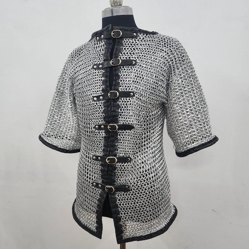May include: A silver chainmail shirt with short sleeves and a black leather closure down the front. The shirt has black trim around the sleeves and the bottom hem. The closure features gold-colored buckles.