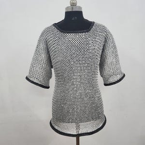 May include: A silver chainmail shirt with short sleeves and a round neckline. The edges of the sleeves and neckline are trimmed with black material. The shirt is displayed on a black mannequin.
