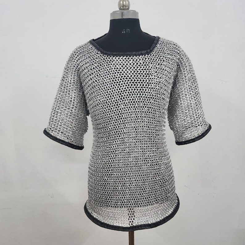 May include: A silver chainmail shirt with short sleeves and a round neckline. The edges of the sleeves and neckline are trimmed with black material. The shirt is displayed on a black mannequin.