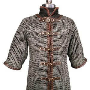 May include: A silver chainmail shirt with brown leather straps and buckles. The shirt has long sleeves and a high neckline.