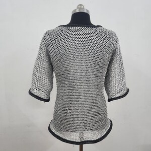 May include: A silver chainmail shirt with short sleeves and a rounded neckline. The edges of the sleeves and neckline are trimmed with black fabric. The shirt is displayed on a mannequin.