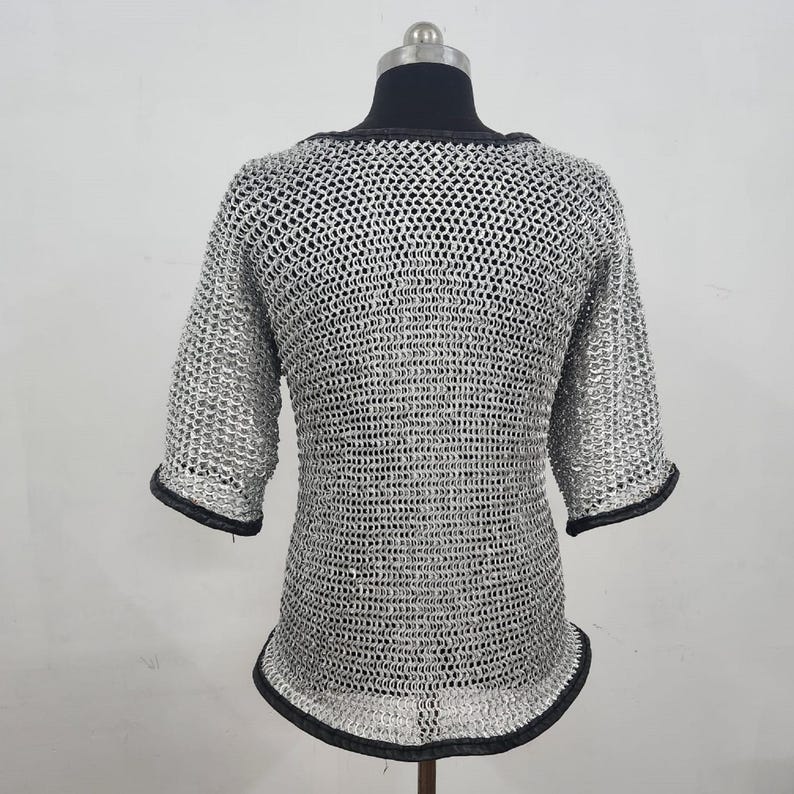 May include: A silver chainmail shirt with short sleeves and a rounded neckline. The edges of the sleeves and neckline are trimmed with black fabric. The shirt is displayed on a mannequin.