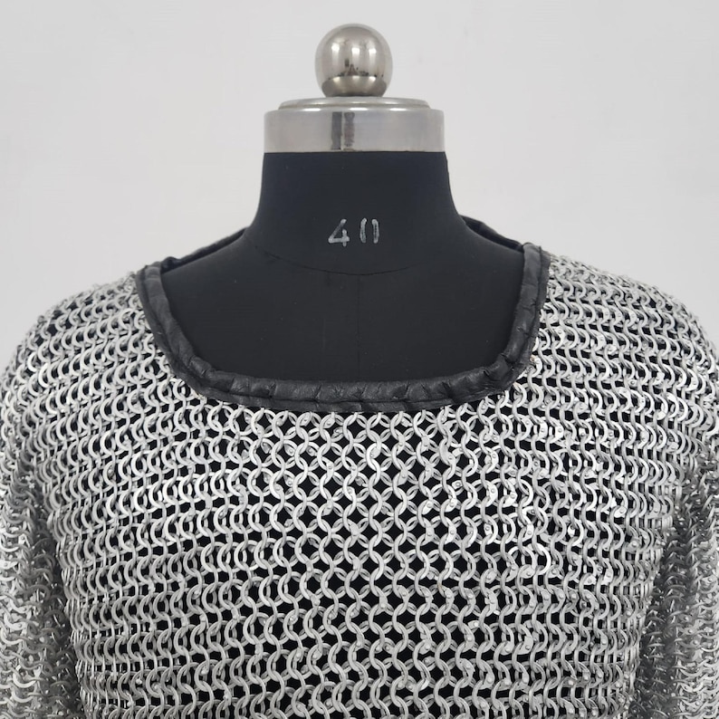 May include: A silver chainmail shirt with a black leather trim around the neckline. The shirt is displayed on a black mannequin with the number 40 visible. The chainmail is made of small, interlocking metal rings.