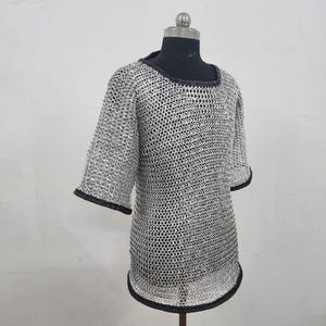 May include: A silver chainmail shirt with short sleeves and a rounded neckline. The edges of the sleeves and neckline are trimmed with black fabric. The shirt is displayed on a mannequin.