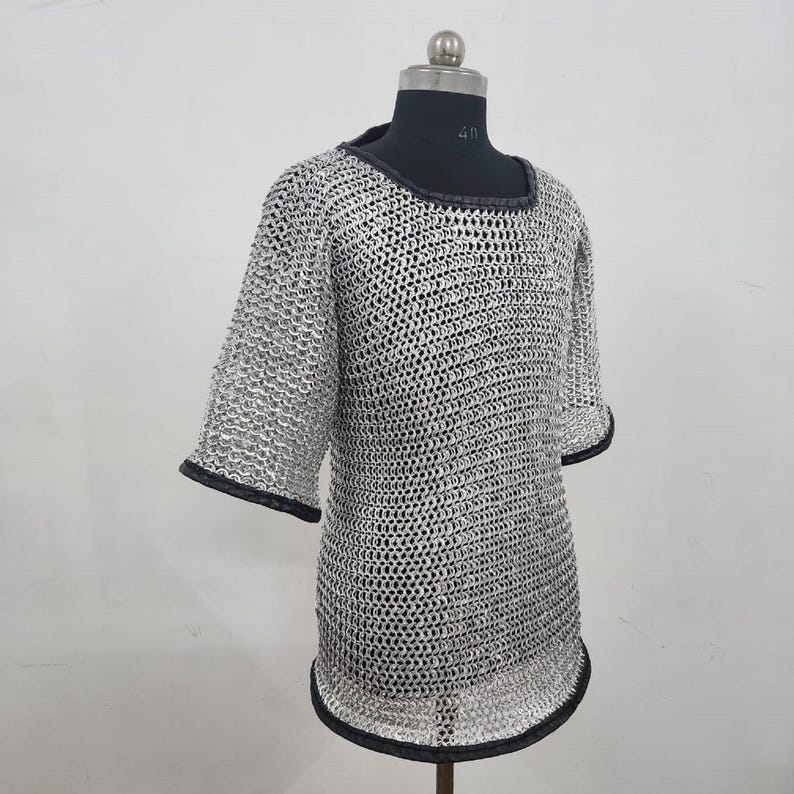 May include: A silver chainmail shirt with short sleeves and a rounded neckline. The edges of the sleeves and neckline are trimmed with black fabric. The shirt is displayed on a mannequin.