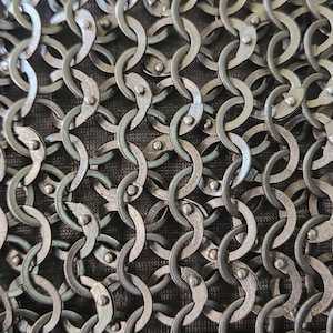 Op de afbeelding: Een close-up van een metalen mali&euml;nkolderstof met in elkaar grijpende cirkelvormige ringen in een donker gunmetal grijze afwerking. Elke individuele ring is vastgezet met een kleine klinknagel, wat een dicht, herhalend gaaspatroon op donkerbruine stof vormt.