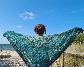 Puede incluir: Una persona sostiene una chal de ganchillo verde azulado con un patrón de encaje en una playa de arena con una barandilla de madera y hierba alta en el fondo.