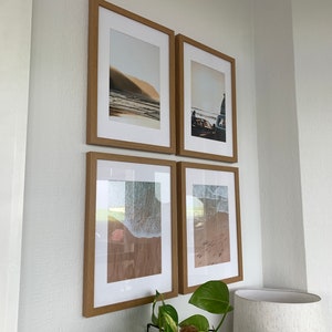 May include: Four framed photographs of beach scenes, arranged in a grid on a white wall. The photos feature a sandy beach, ocean waves, and a vintage car.