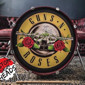 May include: A custom drum head with the Guns N' Roses logo. The design features a yellow centre with the band's name, a gun and roses graphic, and signatures. The drum head is red and black, with a drumstick in front.