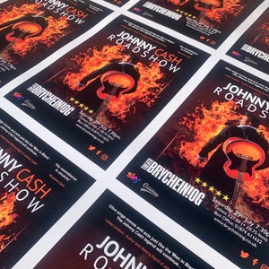 May include: A black and white poster with a fiery background and a silhouette of a man playing a guitar. The poster advertises a Johnny Cash Roadshow at the Brycheiniog Theatre in Wales. The poster includes the date and time of the show, ticket prices, and contact information.