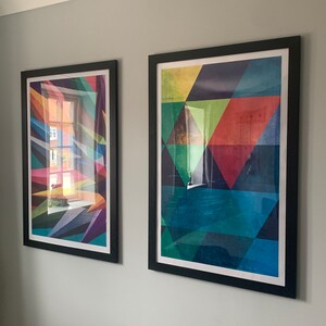 May include: Two framed abstract geometric art prints with black frames. The print on the left has a colorful geometric design with a window reflection. The print on the right has a geometric design with shades of blue, green, red, and yellow.