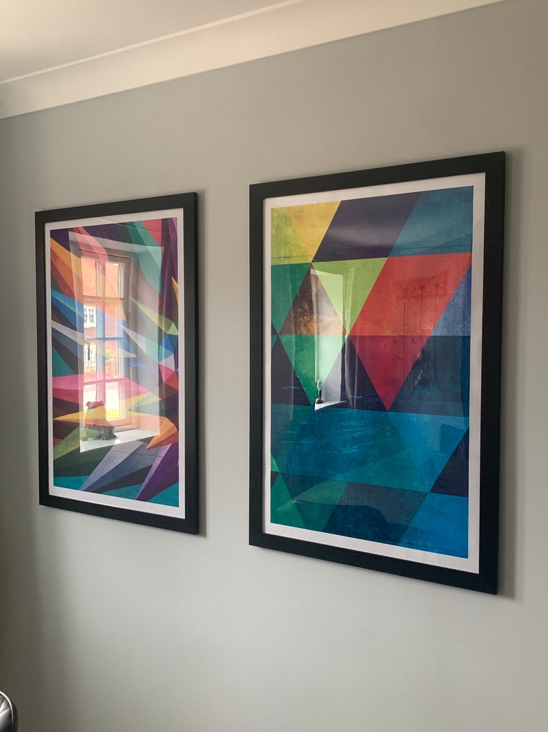 May include: Two framed abstract geometric art prints with black frames. The print on the left has a colorful geometric design with a window reflection. The print on the right has a geometric design with shades of blue, green, red, and yellow.