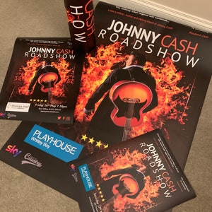 May include: A black and red poster advertising a Johnny Cash Roadshow concert. The poster features a silhouette of Johnny Cash playing a guitar in front of a fiery background. The text "Johnny Cash Roadshow" is printed in white letters on a black background. The poster also includes the dates and times of the concerts.