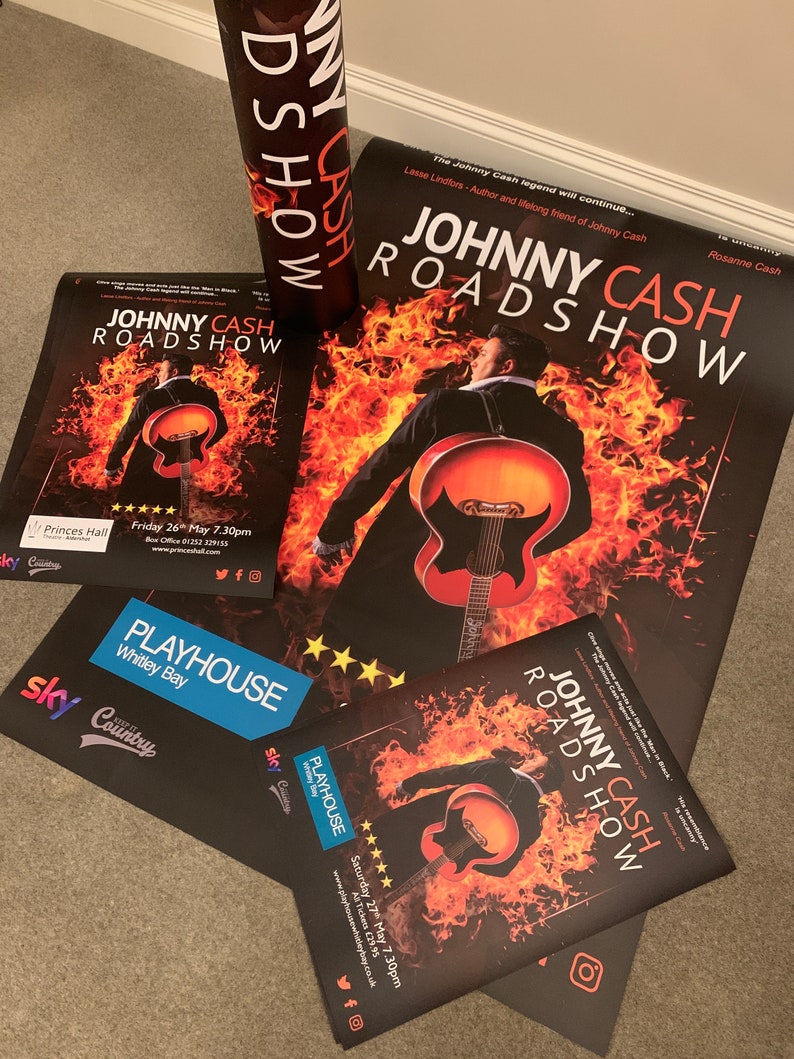 May include: A black and red poster advertising a Johnny Cash Roadshow concert. The poster features a silhouette of Johnny Cash playing a guitar in front of a fiery background. The text "Johnny Cash Roadshow" is printed in white letters on a black background. The poster also includes the dates and times of the concerts.