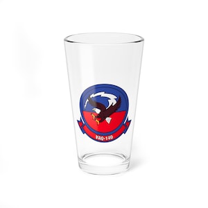 May include: A clear glass pint glass with a blue and red squadron insignia featuring a bald eagle. The insignia reads "VAQ-140".