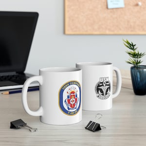 May include: Two white ceramic mugs with a navy blue and gold design. The mug on the left has a navy blue circle with a gold rope border and the text "USNS COMFORT T-AH-20". The mug on the right has a black circle with a white rope border and the text "USN CORPSE" in white.