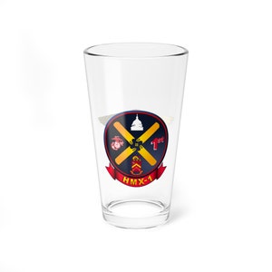 May include: A clear glass pint glass with a blue, red, and yellow graphic of the HMX-1 Marine Helicopter Squadron emblem. The emblem features a crossed pair of yellow propellers, a white Capitol building, and the words "1st" and "HMX-1".