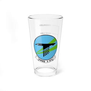 May include: A clear glass pint glass with a black and green squadron insignia. The insignia features a black bird in flight with green wings and a blue background. The text "VAQ 135" is printed below the bird.