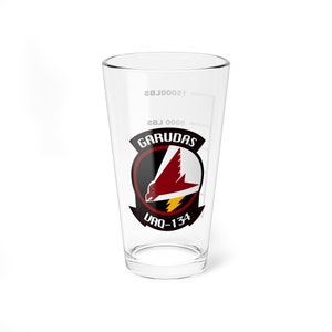 May include: A clear glass pint glass with a white printed design of a squadron patch. The patch features a red bird with a lightning bolt and the text "GARUDAS VAQ-134".