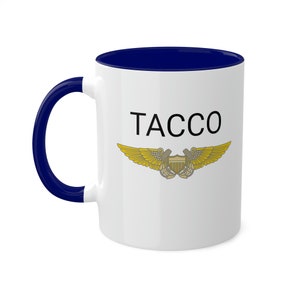 Puede incluir: Taza de café de cerámica blanca con borde y asa azules. La taza presenta un insignia alada dorada con el texto "TACCO" en negro.